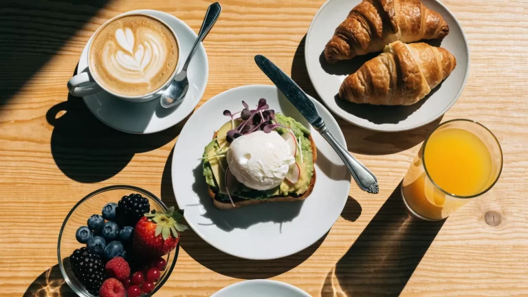 Mesa de brunch con capuchino, tostada de palta con huevo, medialunas y frutas