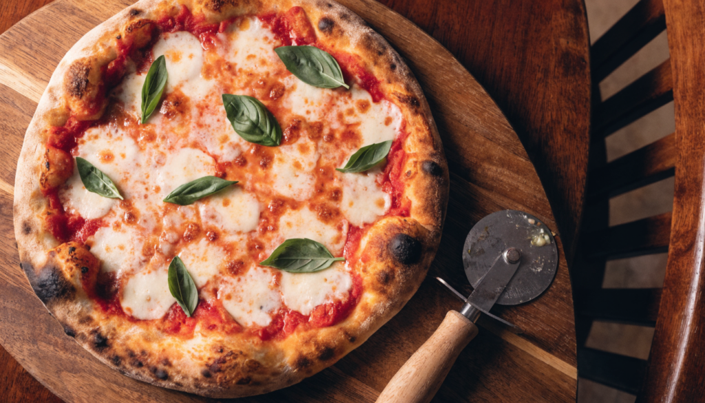 Pizza artesanal recién salida del horno de leña con muzzarella y albahaca sobre tabla de madera. Imagen ilustrativa: no corresponde al local.